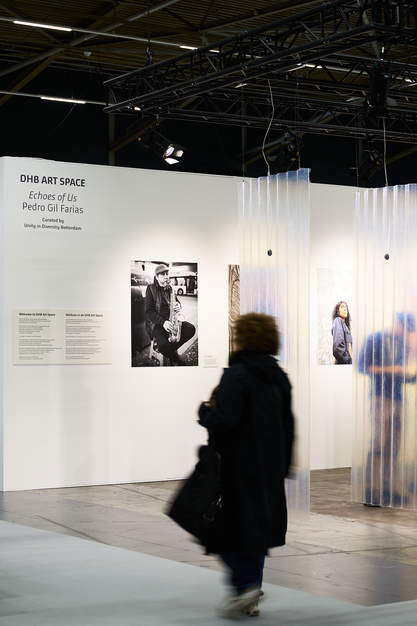 DHB Art Space - Kunstausstellung mit Fotografie Eine Person geht durch den DHB Art Space, eine Kunstausstellung mit Schwarz-Weiß-Fotografien und transparenten Wellenpaneelen. Ein Bild zeigt einen Saxophonisten.